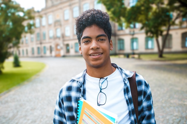 Lo studente afroamericano sorridente con i vetri e con i libri si avvicina all'università. Ritratto di un giovane uomo nero felice in piedi su un'università