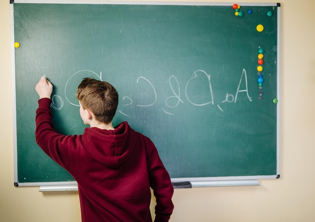 Lo studente di una scuola superiore è in piedi con una felpa con cappuccio scura vicino alla lavagna verde in classe che scrive l'alfabeto Vista dal retro Concetto di istruzione scolastica