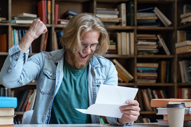 Lo studente felice emozionante che legge la lettera di ammissione si siede alla scrivania della biblioteca