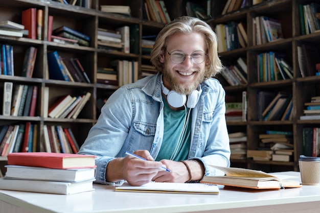 Lo studente felice studia in biblioteca guardando la telecamera e si siede alla scrivania