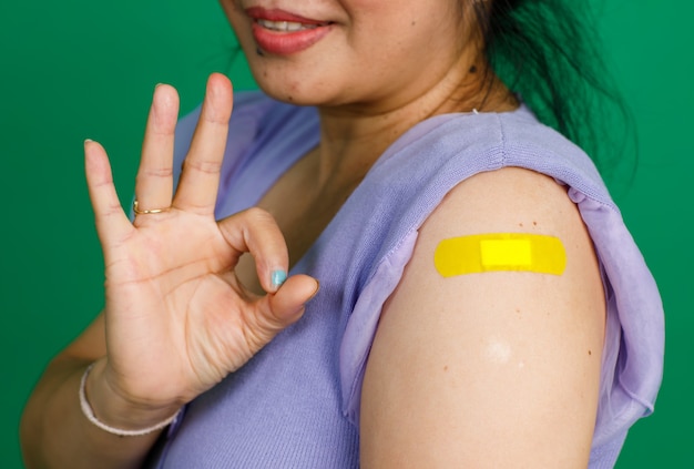 Lo studio fotografico della paziente femmina si siede sorridendo guarda la telecamera mostra il segno della mano ok e la benda di gesso blu sulla spalla dopo aver ricevuto la vaccinazione contro il coronavirus covid 19 su sfondo verde.