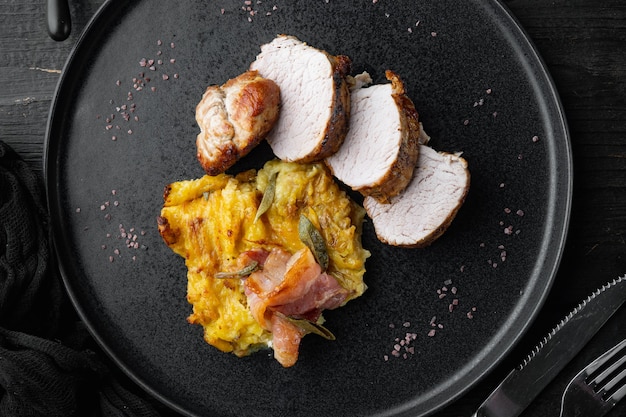 Lonza di filetto di maiale al forno con patate ed erbe aromatiche, su piatto, su fondo di legno nero, vista dall'alto piatta