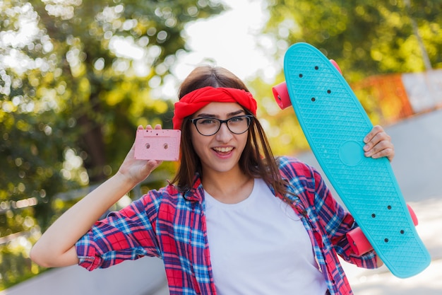 Look giovanile in stile retrò. anni 80. La giovane donna dei pantaloni a vita bassa in vestiti alla moda tiene il pattino e l'audio cassetta mentre posano alla macchina fotografica nello skatepark al giorno soleggiato luminoso