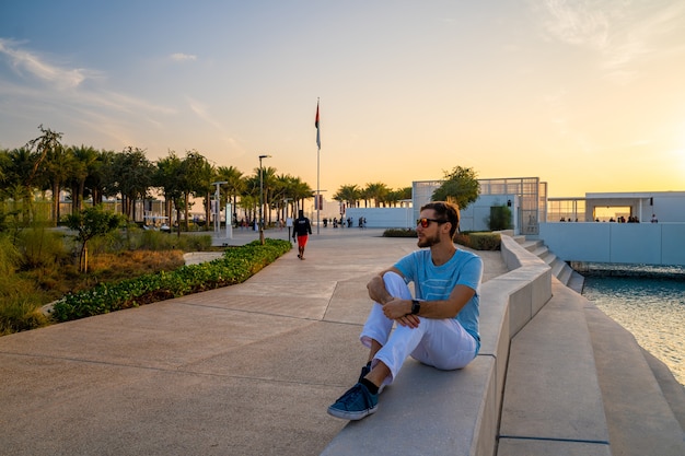 Louvre, Abu Dhabi, Emirati Arabi Uniti - 10 maggio 2020. Giovane seduto vicino al louvre ad Abu Dhabi. Bellissimo edificio al tramonto.