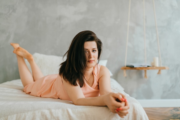 Luiyng bello del ritratto della donna del brunette sul letto bianco a casa in mattina.