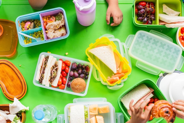 Lunchbox colorate con panini frutta e spuntini su un tavolo verde Bambini39 mani che raggiungono il cibo Lunchbox salutari Lunchbox vivaci Lunchbox gustosi Lunchbox Bambini che pranzano a scuola