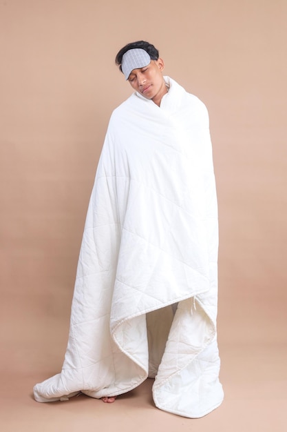 Lunghezza completa di giovane uomo indossare pigiama marmellata e maschera da letto a casa avvolto con coperta piumetto isolato o