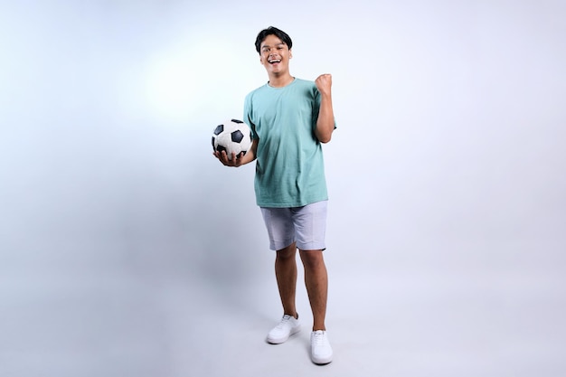 Lunghezza completa di un giovane asiatico che tiene la palla da calcio e stringe il pugno per festeggiare la vittoria