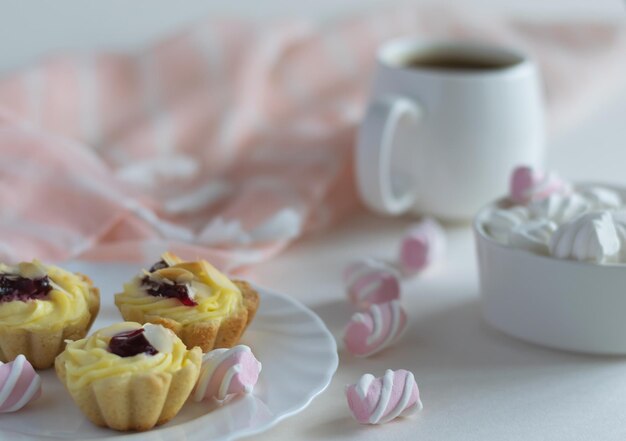 Lussureggianti pan di spagna alla frutta con mandorle su un piattino bianco, mini marshmallow in una tazza di porcellana e una tazza di caffè forte. Foto ravvicinata su sfondo chiaro. Messa a fuoco selettiva.