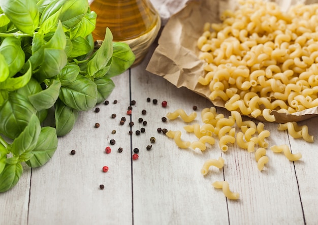 Maccheroni gomiti la classica pasta cruda in carta marrone su fondo in legno chiaro con basilico e olio.