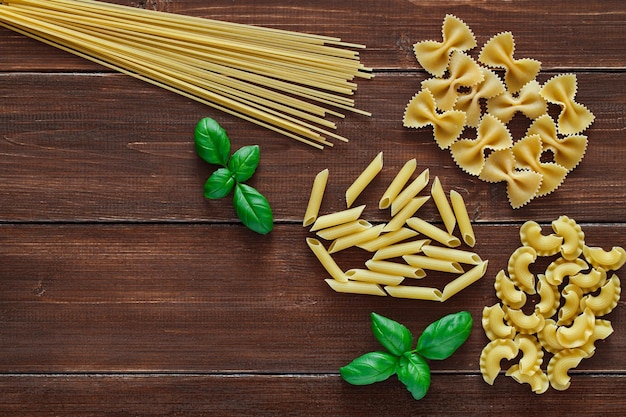 Maccheroni spaghetti crudi farfalle penne rigate cresta di gallo foglia di basilico su tavole di legno marrone sfondo scuro vista dall'alto spazio per copiare il testo