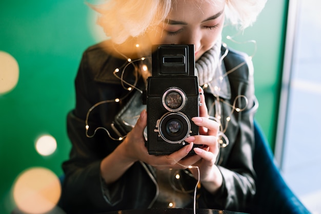 Macchina da presa d'annata della giovane tenuta femminile asiatica bionda con le luci leggiadramente e il bokeh intorno, fuoco selettivo