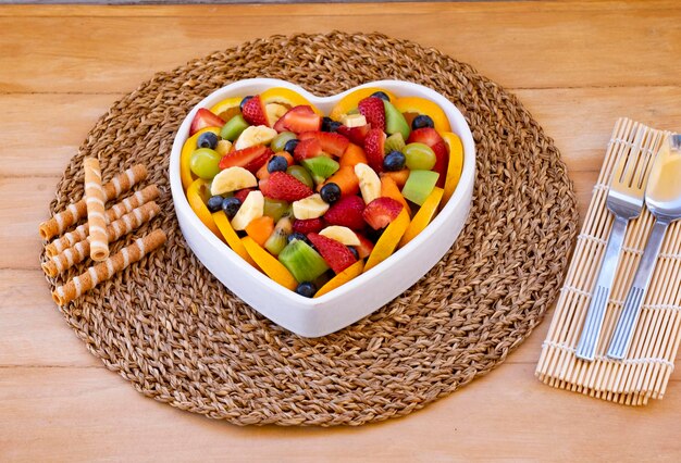 Macedonia di frutta fresca in un piatto a forma di cuore. Tavolo rustico in legno, biscotti. Mangiare sano