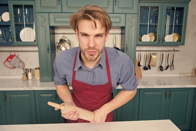 Macho con faccia seria in grembiule da chef rosso con supporto per mattarello al tavolo in cucina Cuocere cuocere cucina culinaria Concetto di cibo domestico