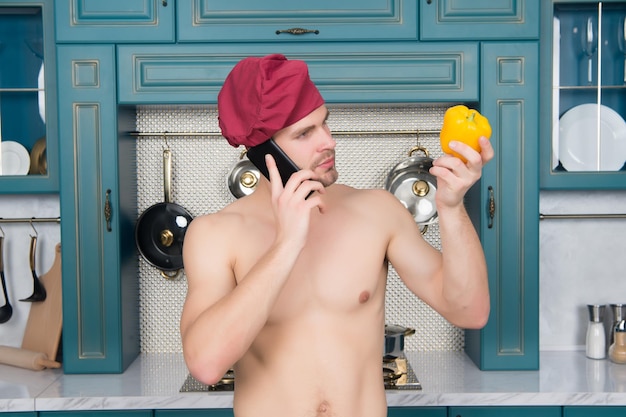 Macho con il torso sexy in cappello da chef parla al cellulare e guarda la verdura di peperone giallo in cucina.