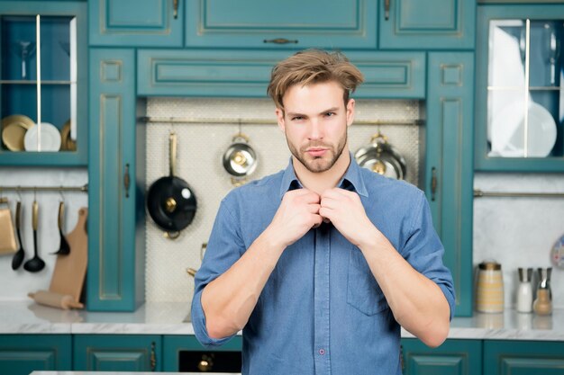 Macho in camicia blu fissa il colletto sulla cucina. Preparazione del cibo, cucina, concetto di cucina. Moda, stile, tendenza.