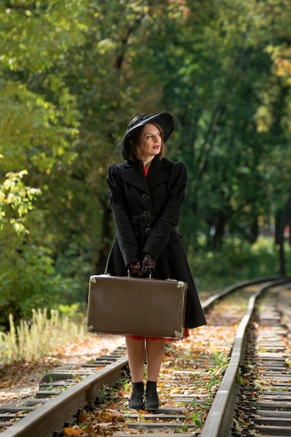 Madame in abiti di stile anni '40 distoglie misteriosamente lo sguardo La donna con cappello e cappotto neri tiene una valigia retrò Cornice verticale