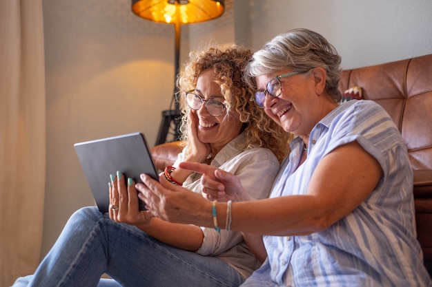 Madre anziana con una bellissima figlia adulta a casa che sorride insieme guardando qualcosa di divertente sul dispositivo digitale