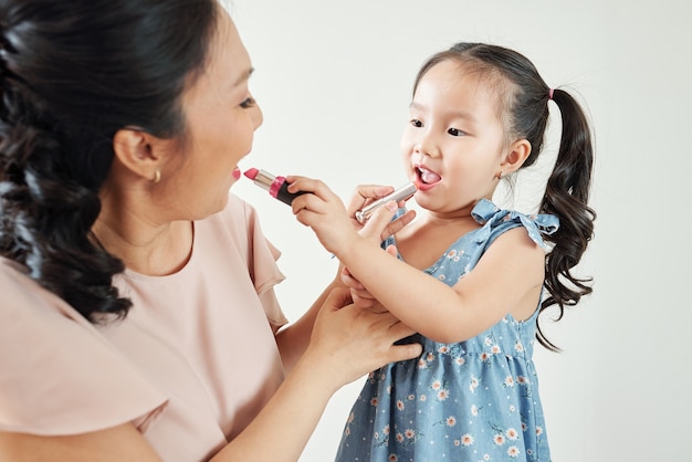 Madre asiatica allegra e figlia che applicano i rossetti sulle labbra l'una dell'altra quando si prepara la mattina