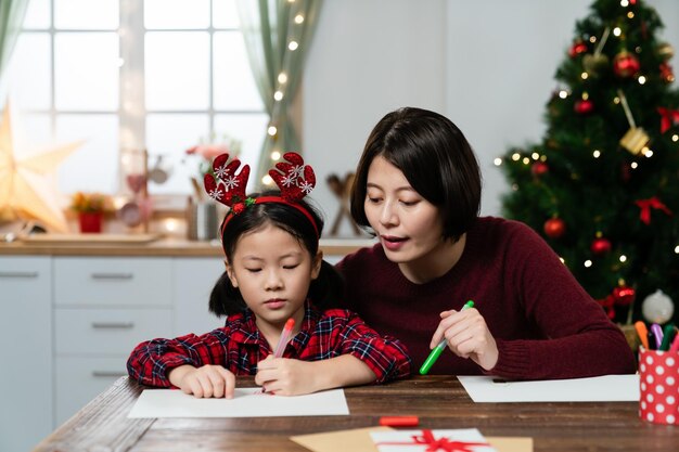 madre asiatica che aiuta la sua giovane figlia a fare la lista dei desideri di regali per Natale a tavola in una stanza luminosa con decorazioni natalizie