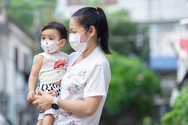 Madre asiatica che indossa una maschera bianca che tiene in braccio la sua piccola bambina che è sua figlia indossa una maschera facciale, guardano fuori, concetto COVID-19