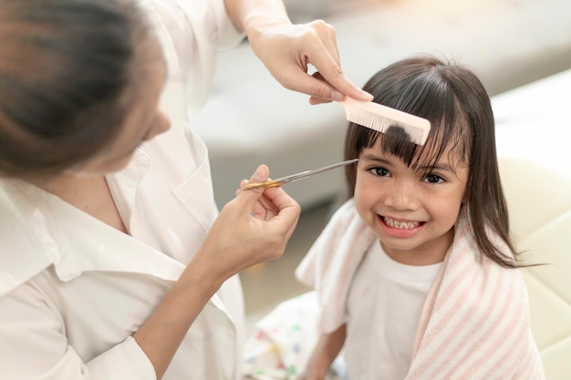 Madre asiatica che taglia i capelli a sua figlia nel soggiorno di casa mentre resta a casa al sicuro dal Coronavirus Covid19 durante il blocco Concetto di autoquarantena e distanza sociale