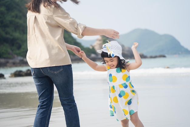 Madre asiatica e sua figlia sorridenti che ridono ballano insieme sulla spiaggia vista mare blu cielo e sfondo di montagna durante il giorno mare calmo durante le vacanze del fine settimana Viaggi rilassati sull'isola