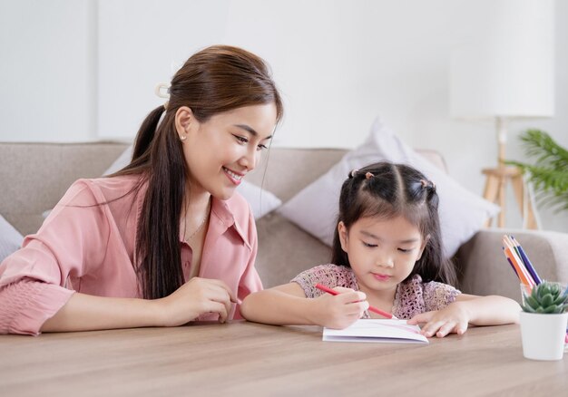 Madre asiatica la sua piccola figlia nel soleggiato salotto accogliente mamma insegna alla ragazza a dipingere carta e matite