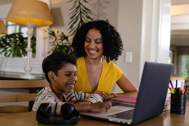 Madre bi-razziale sorridente che guarda suo figlio che studia online sul portatile a casa