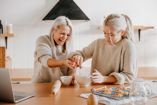 Madre bionda di mezza età e figlia adolescente in cucina guardando la ricetta o il programma televisivo sul laptop e cucinando il concetto naturale di stile di vita