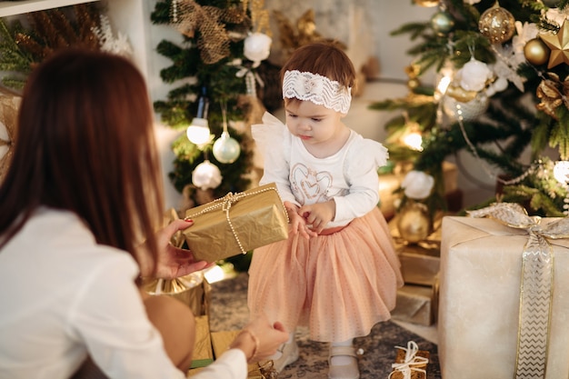 Madre castana che dà il regalo di Natale alla piccola figlia adorabile in gonna rosa, maglione bianco e fascia