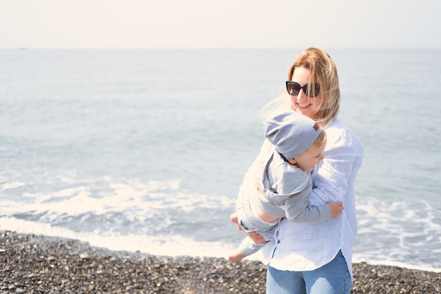 Madre che abbraccia il suo piccolo figlio che ride, famiglia divertendosi sulla riva del mare in giornata di sole.