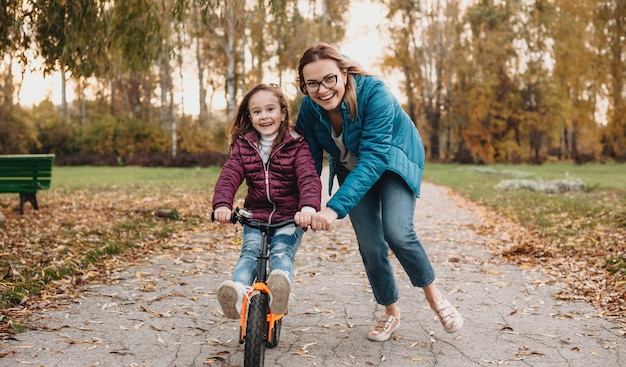 madre che guarda attraverso gli occhiali sta aiutando la figlia ad andare in bicicletta durante una passeggiata autunnale