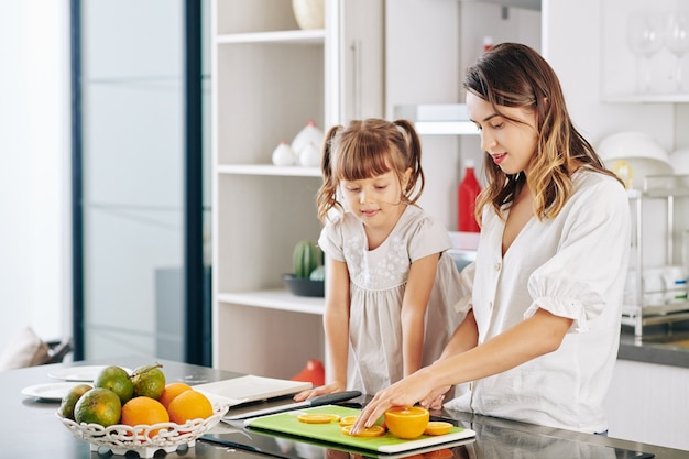 Madre che mostra al suo piccolo dauhter come tagliare l'arancia matura a fettine sottili al bancone della cucina