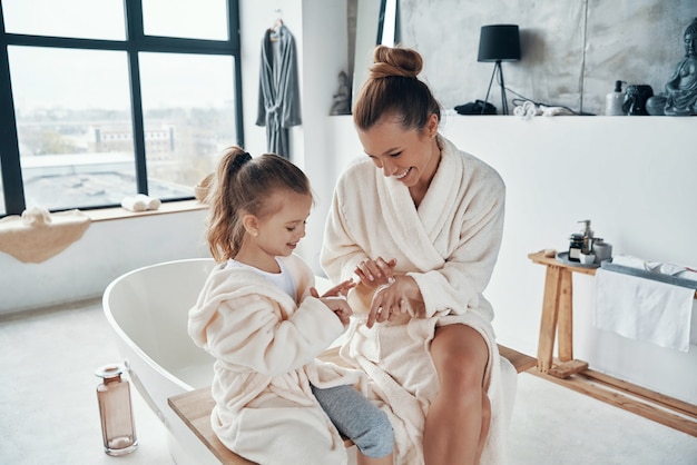 Madre con figlia in accappatoio che sorride e usa la crema per le mani mentre fa la routine mattutina