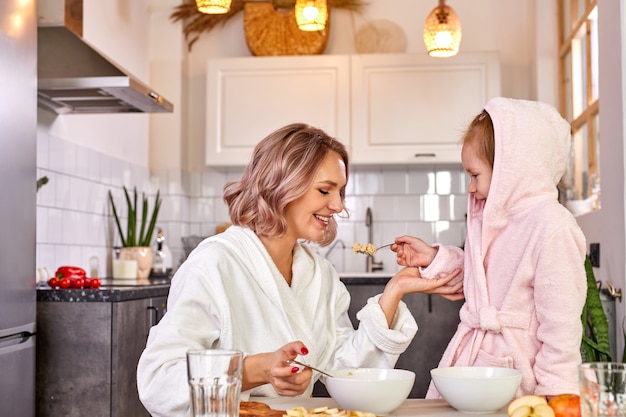 Madre con la ragazza del bambino che mangia il porridge gustoso per colazione, la figlia sta trattando la madre con il cucchiaio, nella cucina leggera a casa