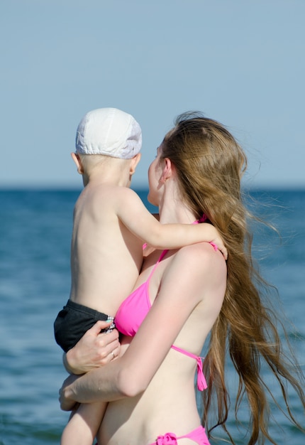 Madre con suo figlio in braccio in costume da bagno in piedi sulla spiaggia e guardare il mare