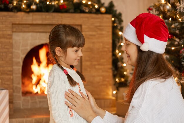 Madre di famiglia felice e piccola figlia che giocano alla vigilia di Natale, in posa vicino al camino, mamma che dice alla sua bambina qualcosa di interessante