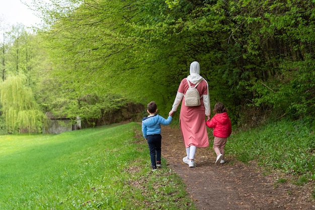 Madre di famiglia musulmana felice in hijab che tiene le mani dei bambini mentre cammina attraverso la foresta del sentiero