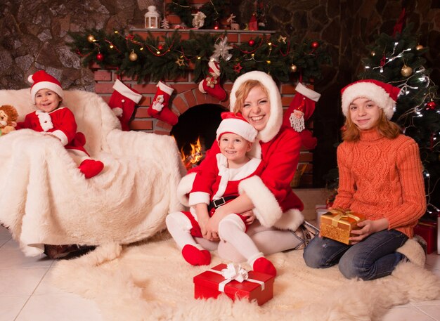 Madre e bambini sono seduti vicino al camino e all'albero di Natale con scatole regalo. Fratello e sorella in abiti da Babbo Natale