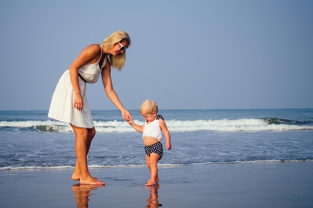 Madre e bambino che insegnano alla sua dolce figlia a fare i suoi primi passi su una spiaggia sabbiosa del mare tropicale. primo passo sulla spiaggia sabbiosa vicino all'oceano nella soleggiata mattina. Concetto di festa della mamma