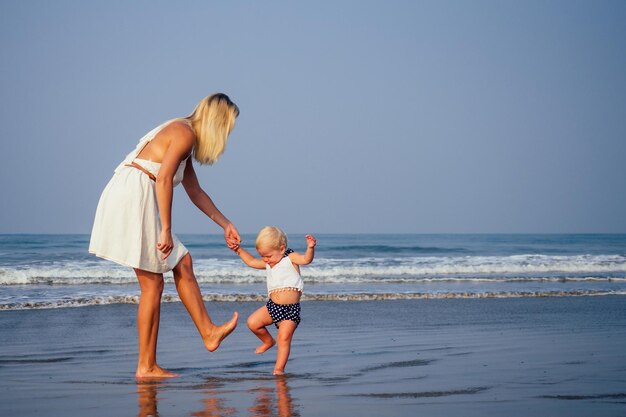 Madre e bambino giovane donna insegnando alla sua dolce figlia del bambino a camminare i suoi primi passi su una spiaggia sabbiosa del mare tropicale primo passo sulla spiaggia sabbiosa vicino all'oceano nella soleggiata mattina Concetto di festa della mamma