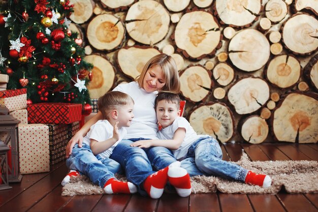 Madre e due ragazzi con il cappello di Natale sono seduti vicino all'albero di Capodanno sullo sfondo di tagli di legno.