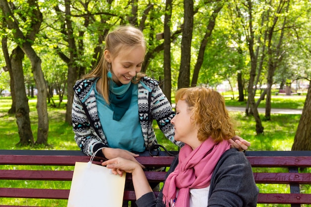 Madre e figlia adolescente con la borsa della spesa che sorridono e parlano su una panchina del parco