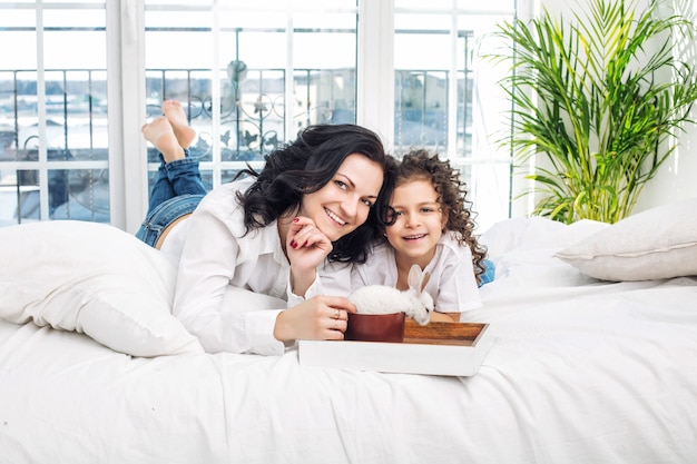 Madre e figlia carine belle e felici a letto con coniglio bianco sul vassoio in camera da letto