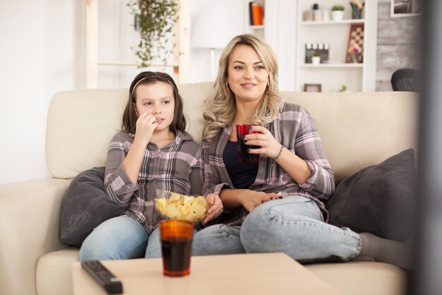 Madre e figlia che guardano un film seduti sul divano in una giornata pigra mangiando patatine e bevendo soda.