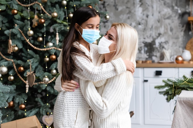 madre e figlia che indossano maschere mediche sotto l'albero di natale durante la celebrazione domestica.