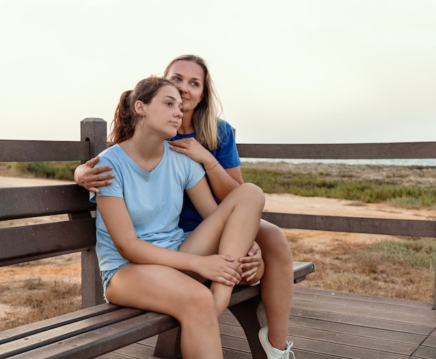 Madre e figlia che si siedono insieme su una panca di legno al tramonto. Sorridente donna di mezza età che abbraccia le anime adolescenti. Modello di t-shirt