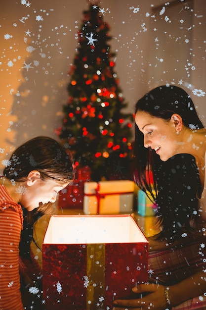 Madre e figlia festose che aprono un regalo di natale incandescente contro la caduta della neve