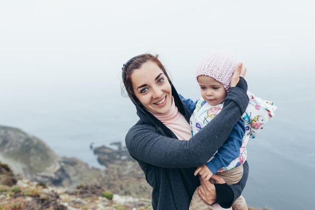 Madre e figlia in piedi rock ocean beach stagione autunnale. Famiglia caucasica con un bambino che viaggiano insieme. Mamma che tiene in mano una bambina di 2 anni e sorride sullo sfondo del paesaggio marino